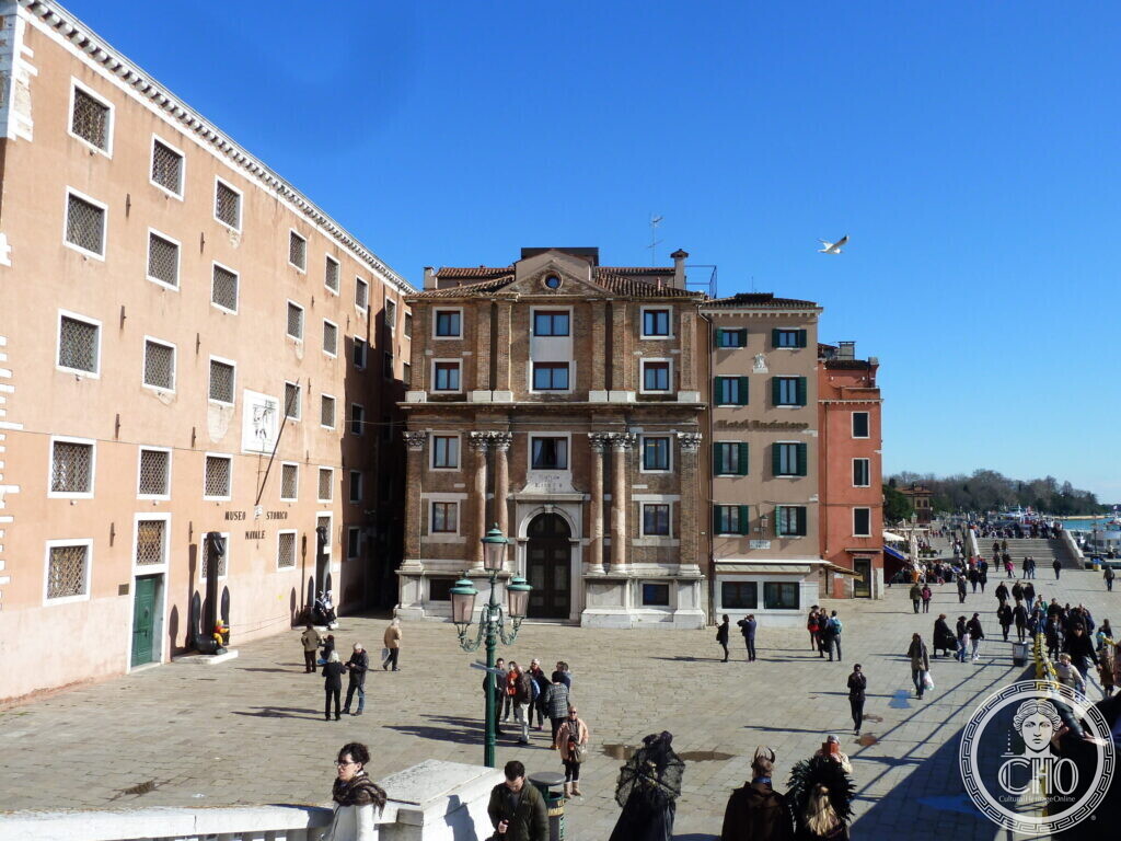 A wide paved square with the long pink building of the naval museum on the left and the brick and stone front of San Biagio in the centre