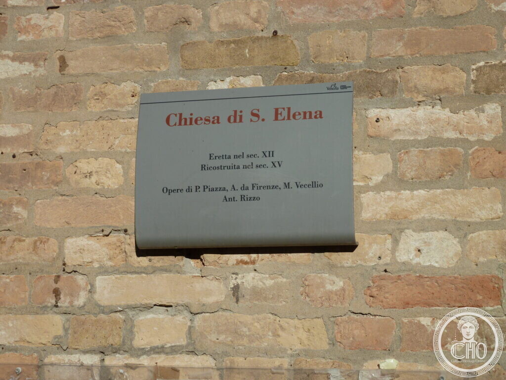 A grey metal plaque on a stone wall headed Chiesa di S. Elena, with its dates and artists