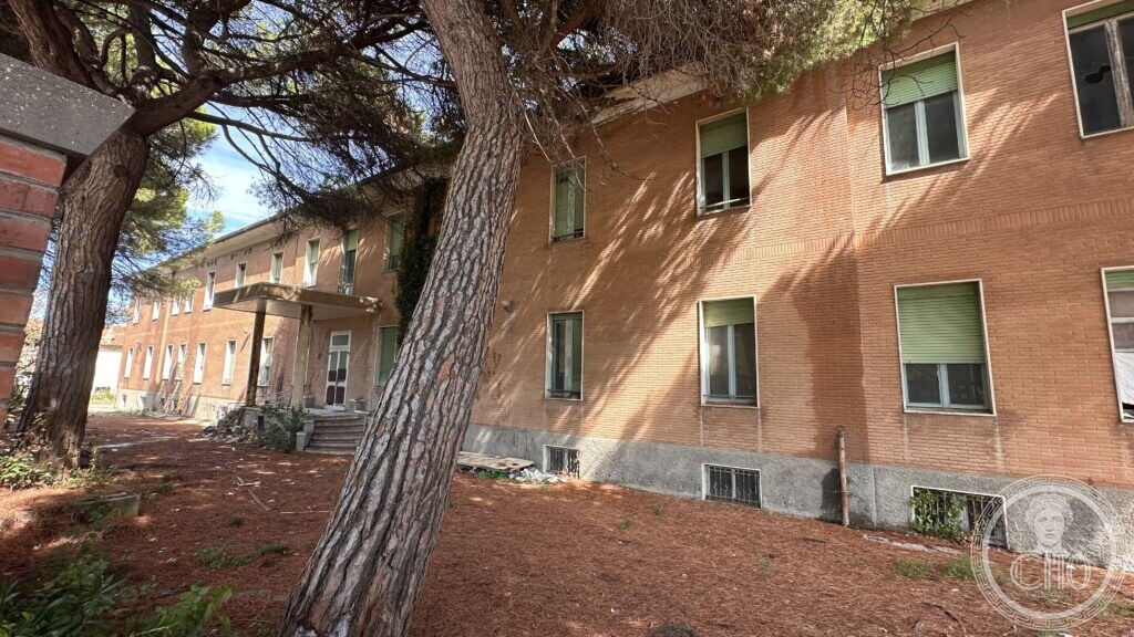 Side entrance of the colony building with a covered porch, ivy climbing the brick, and broken window shutters