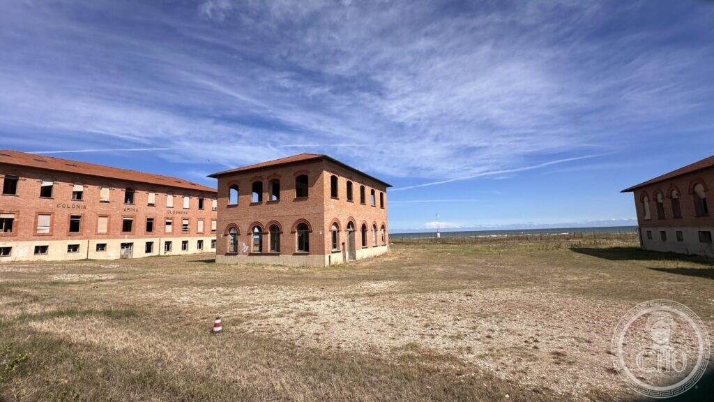 An empty gravel courtyard between brick pavilions, with the Adriatic on the horizon