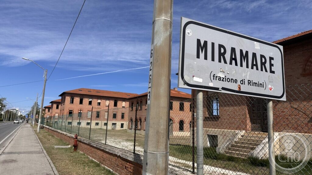 A road sign reading MIRAMARE, frazione di Rimini, with the brick colony behind it