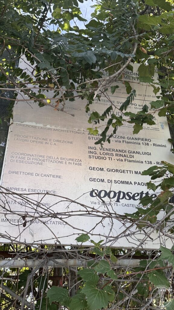 A weathered site notice board for building works, overgrown by brambles, with the names of the designers and the contractor still legible