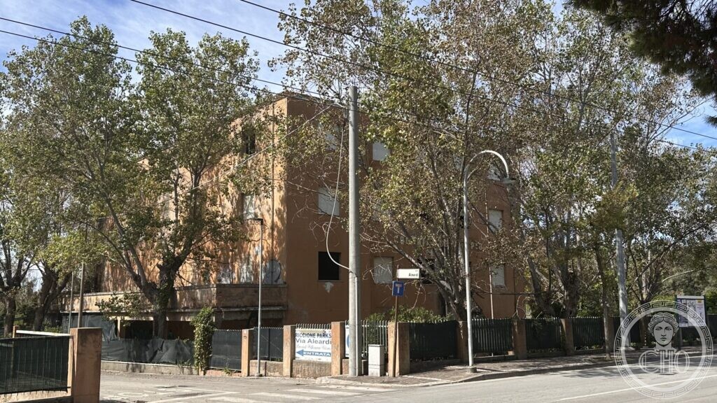 A plain three-storey ochre block at a street corner, behind trees and a green fence