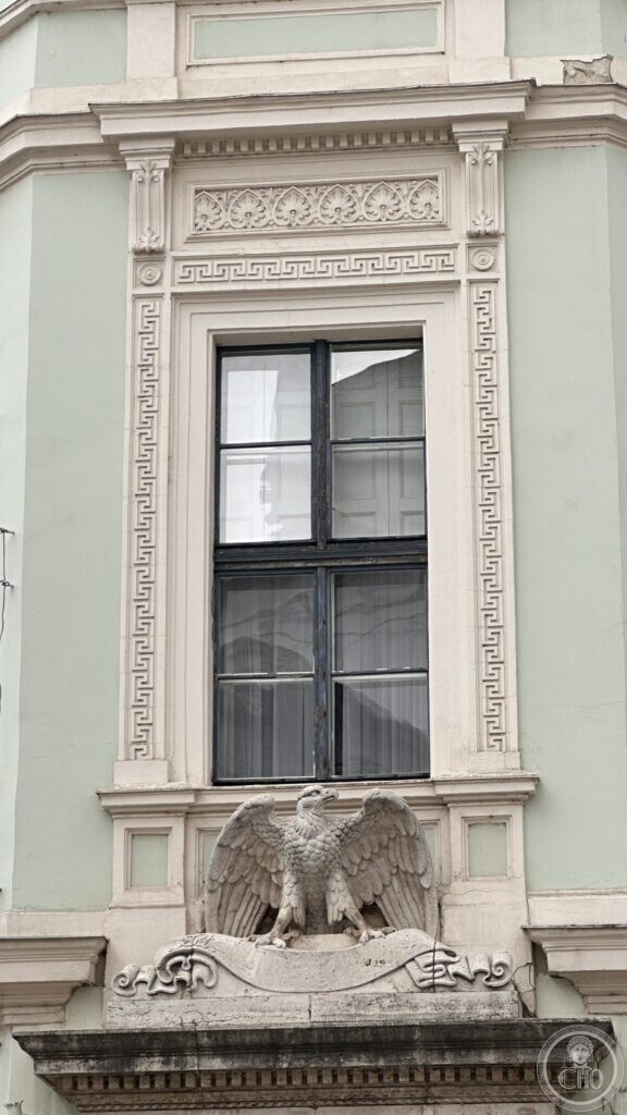 The corner bay of the building, with a tall window framed by bands of Greek key ornament and a carved eagle on a scroll below it