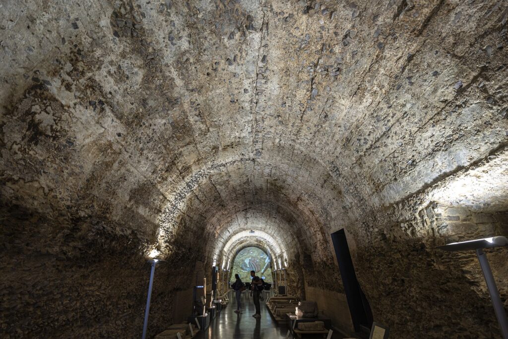 Interior of the Ergástula of Astorga, a long Roman barrel-vaulted gallery of rough concrete lit from below, with museum display stands along the floor and two visitors at the far end