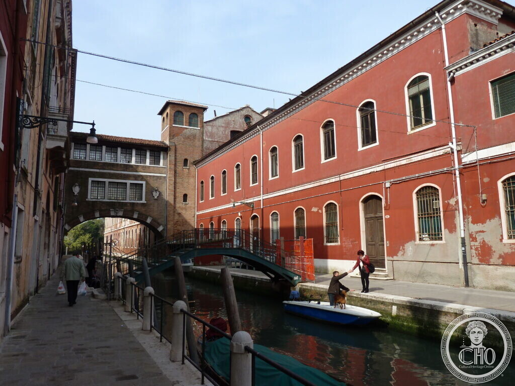 Red-plastered industrial buildings of the former Manifattura Tabacchi along a canal, with a footbridge and moored boat, Venice