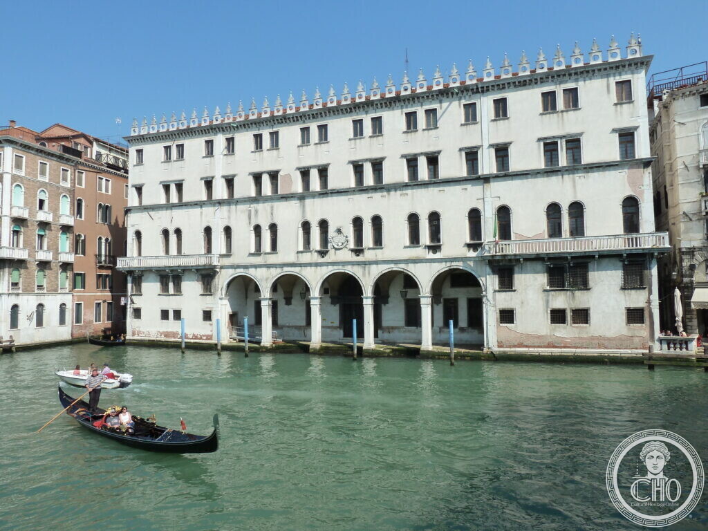 The Fondaco dei Tedeschi seen from the Grand Canal in 2013, its rendering weathered and grey, with the five-arched water loggia and the crenellated roofline, a gondola passing in front