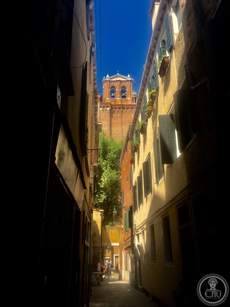Sunlight and shadow down a narrow Venetian calle framing the towering brick bell tower of San Lazzaro dei Mendicanti in Castello Venice