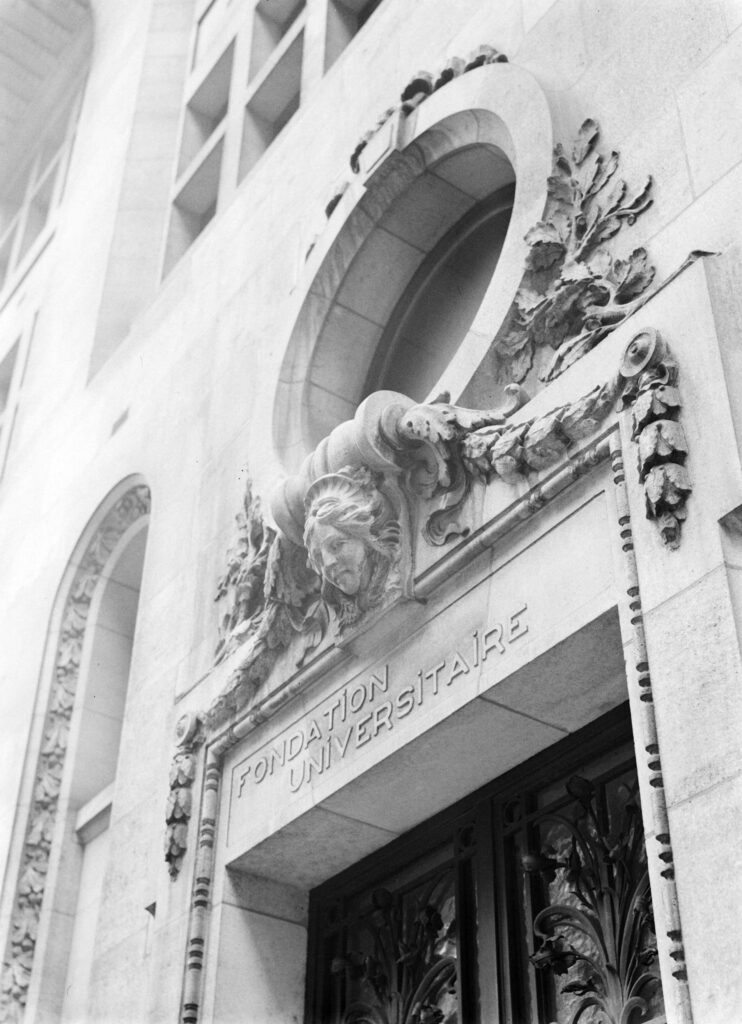 Carved stone doorway with the words Fondation Universitaire, an oval window above, a female mask and oak-leaf garlands, photographed in 1935