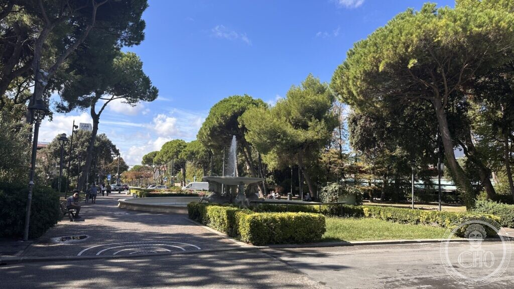 The fountain seen from a path under stone pines, with hedges and benches around it