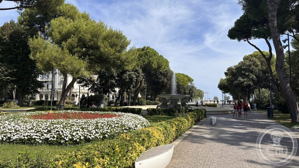 The park with flower beds, the fountain playing in the middle distance and a large white hotel behind the trees