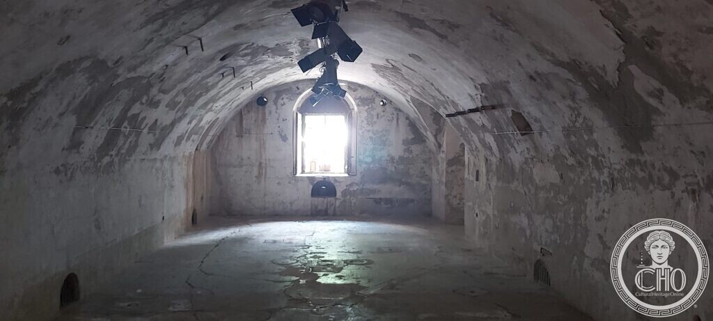 A long empty room with a plastered barrel vault and peeling walls, lit by a single arched window at the far end, lights hanging from the ceiling