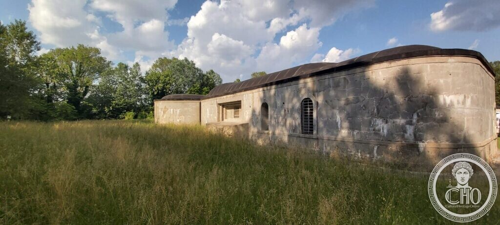 The curved grey concrete side of a low fort with a small window, an arched door and a barred arched opening, tall grass in front