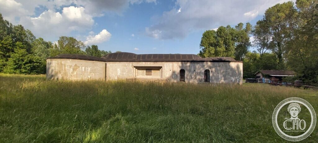 A long low grey concrete fort with a curved front and a dark flat roof, standing in a meadow of tall grass with trees behind