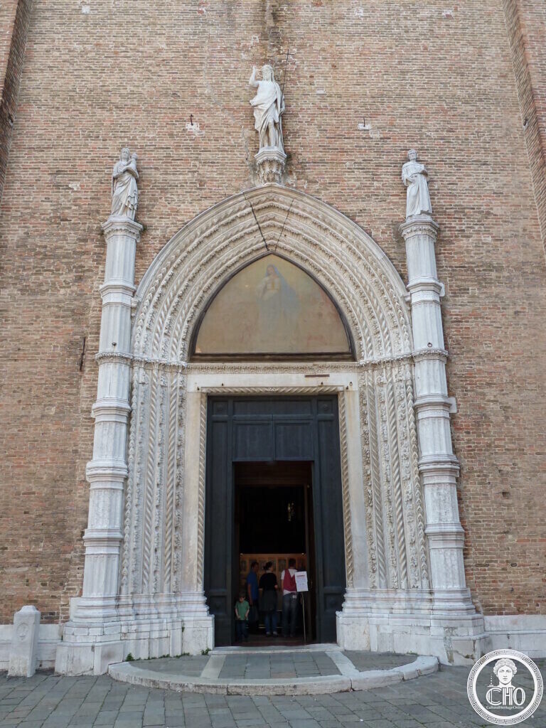 The pointed marble portal of the Frari basilica in Venice, its arch carved in rope mouldings, a faded fresco in the lunette, a statue of the risen Christ above and two saints on the flanking columns