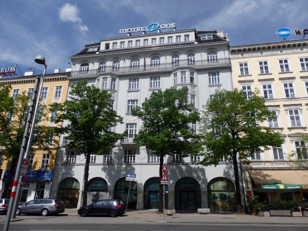 White Secession-era commercial building with a polygonal corner oriel, an iron balcony with arched windows near the top and large arched openings on the ground floor, Friedrichstraße, Vienna