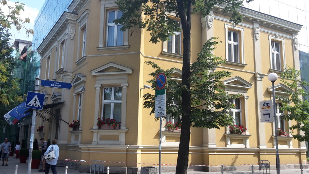 The ochre neoclassical facade of the Georgov House at the corner of Shipka and Krakra streets, Sofia, with pedimented windows and pilasters, and a modern glass building rising behind it