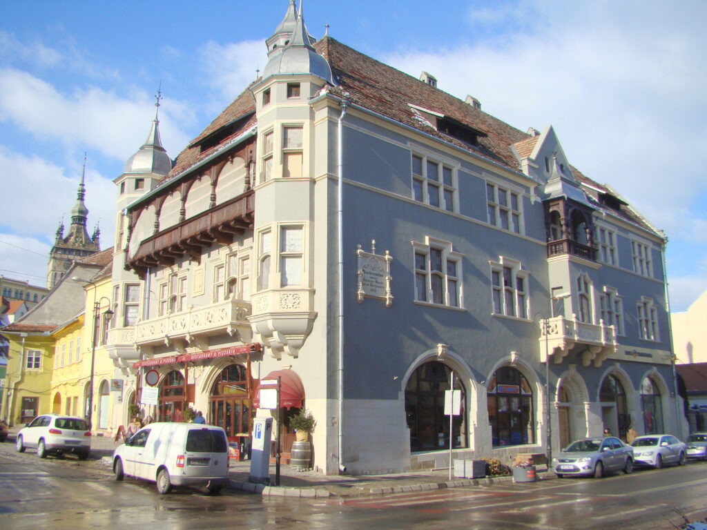 The Guilders' Association House on Hermann Oberth Square, Sighișoara: grey-blue and cream facade with corner turrets, a timber gallery under the roof and arched shop windows, the citadel clock tower behind