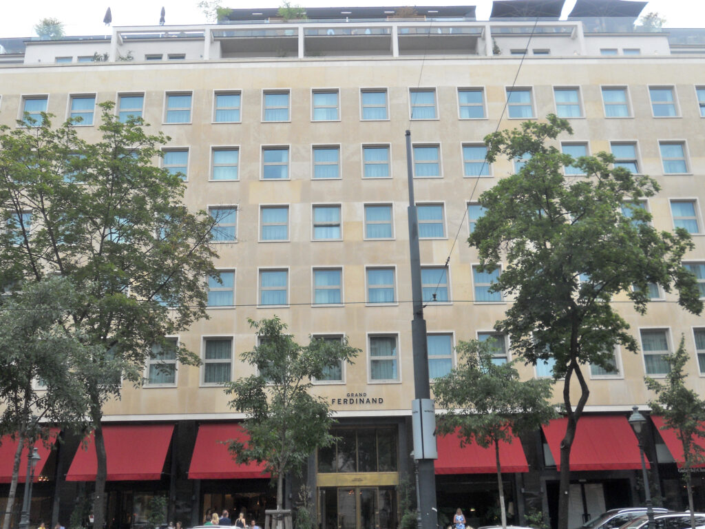 Long multi-storey front clad in pale stone with rows of square windows, the name GRAND FERDINAND above a brass-framed entrance and red awnings along the ground floor, Schubertring, Vienna