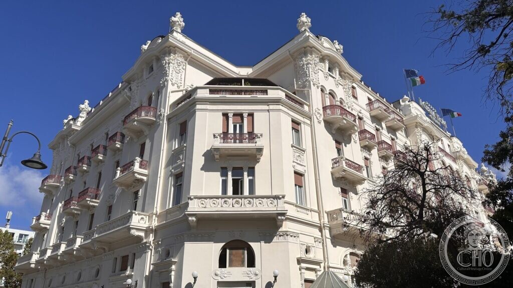 The white Liberty facade of the Grand Hotel of Rimini seen from the street corner, with balconies and roof urns