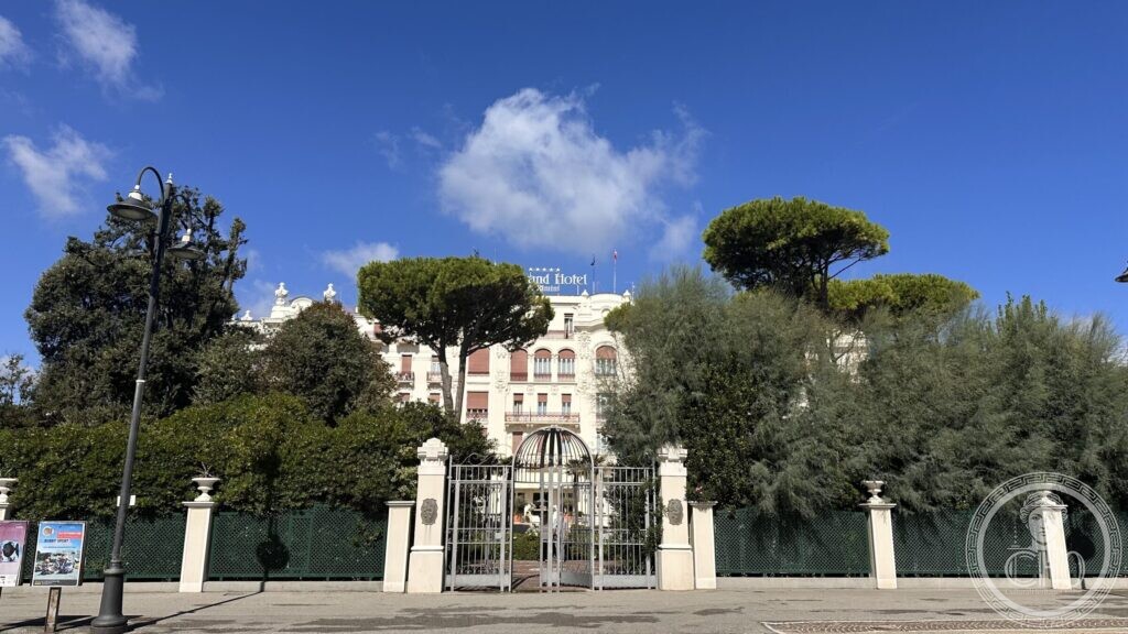A white gate with stone piers and a lantern, with the hotel and pines beyond