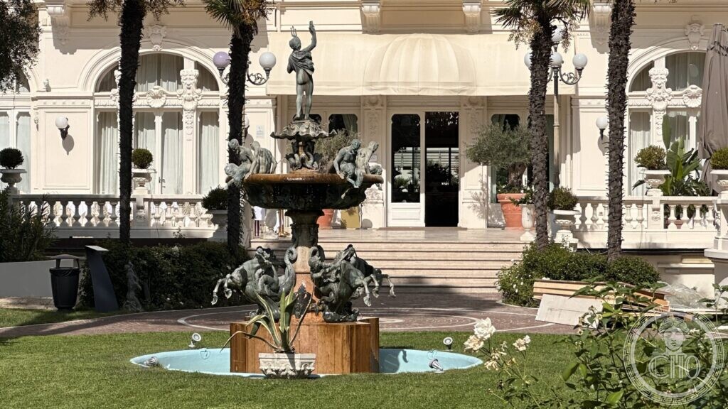 A bronze fountain with putti and a figure on top, in a pool in front of the hotel veranda