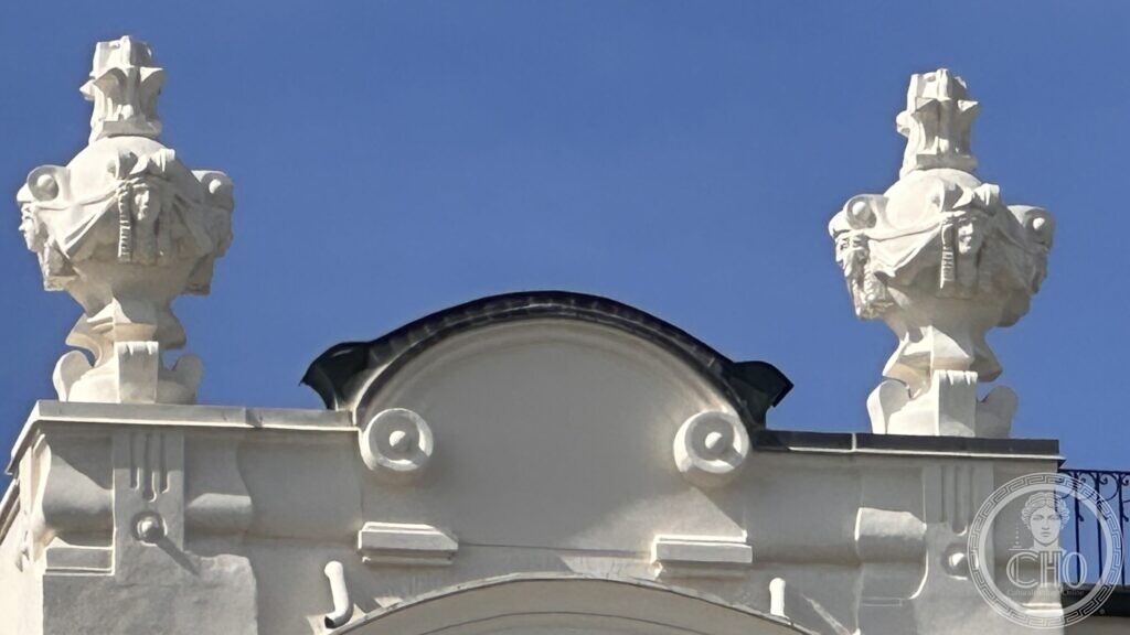 Two carved stone urns standing against the sky above the curved pediment of the hotel