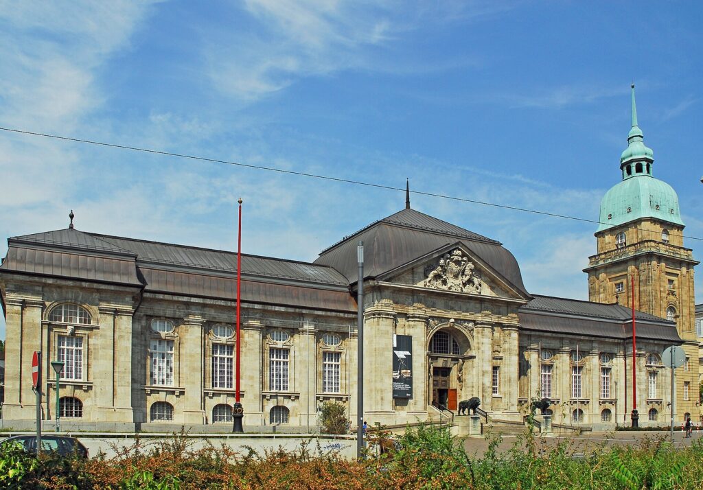 The Hessisches Landesmuseum in Darmstadt: a long two-storey stone building with a pedimented entrance pavilion, bronze lions at the steps and, at the right, a stone tower with a green copper dome and spire