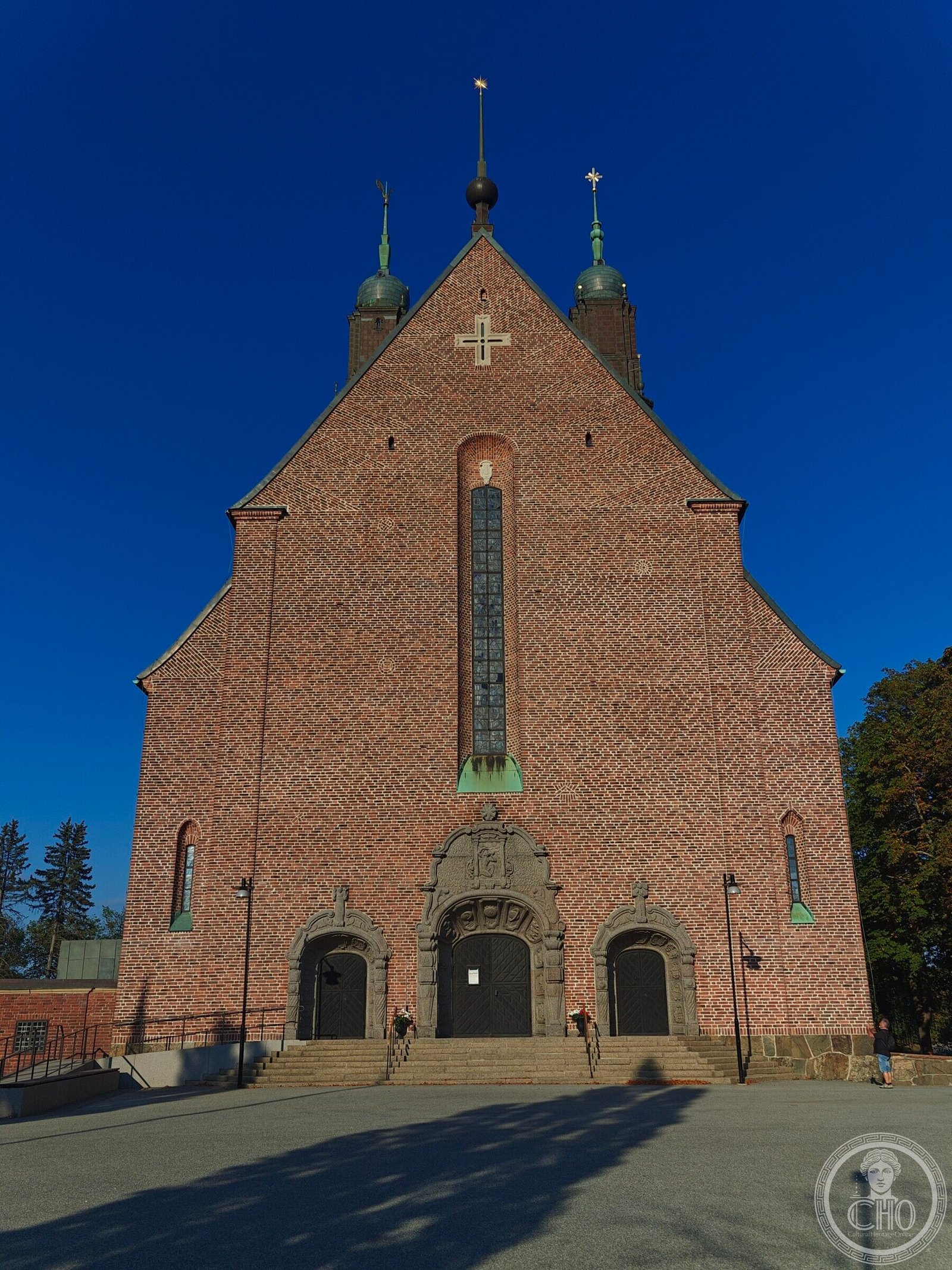 The brick west front of Högalidskyrkan, with a tall stained-glass window, a stone cross at the gable peak, and three carved granite doorways