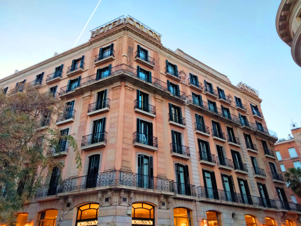 Corner building in salmon stucco with iron balconies, decorated pilasters and HOTEL COLÓN letters on the roof, facing Barcelona Cathedral
