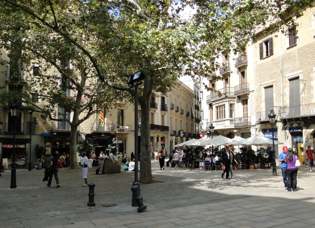 Plaça de Sant Josep Oriol in Barcelona shaded by plane trees; on the left a corner house with stone-patterned stucco and iron balconies, the blue awning of the Bar del Pi at street level and the seated statue of Àngel Guimerà in front