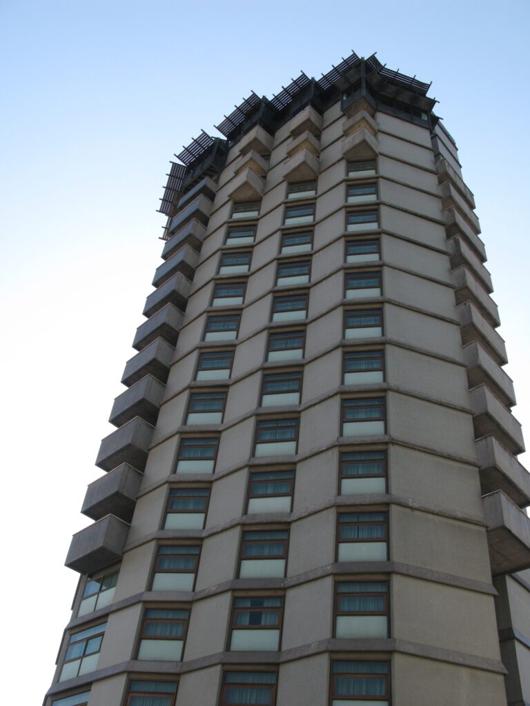 The Hotel D. Henrique tower in Porto seen from below: a many-sided concrete shaft banded at every floor, with staggered projecting window boxes and a dark metal crown against a pale sky