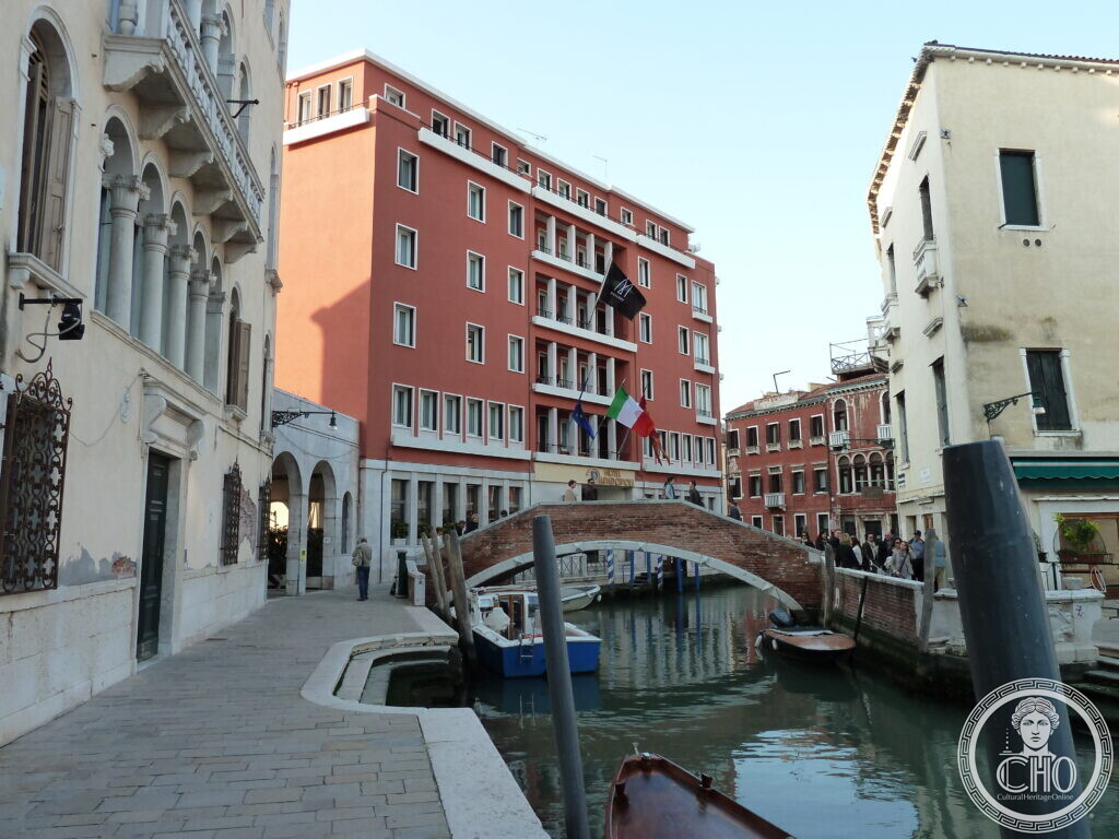 Looking along the Rio dei Tolentini in Venice towards the bridge and the red facade of the Hotel Papadopoli, with flags on the corner and boats moored along the quay