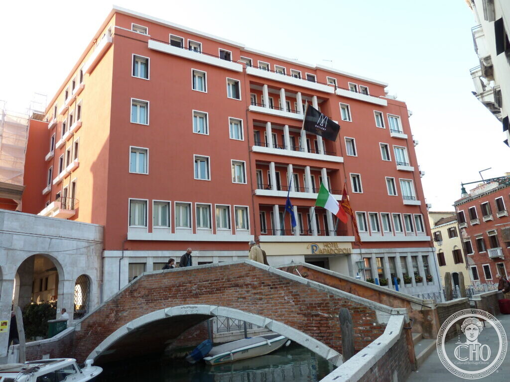 The red-rendered block of the Hotel Papadopoli in Venice with its recessed balconies and the gilded hotel sign, seen from the brick bridge over the canal in front of it