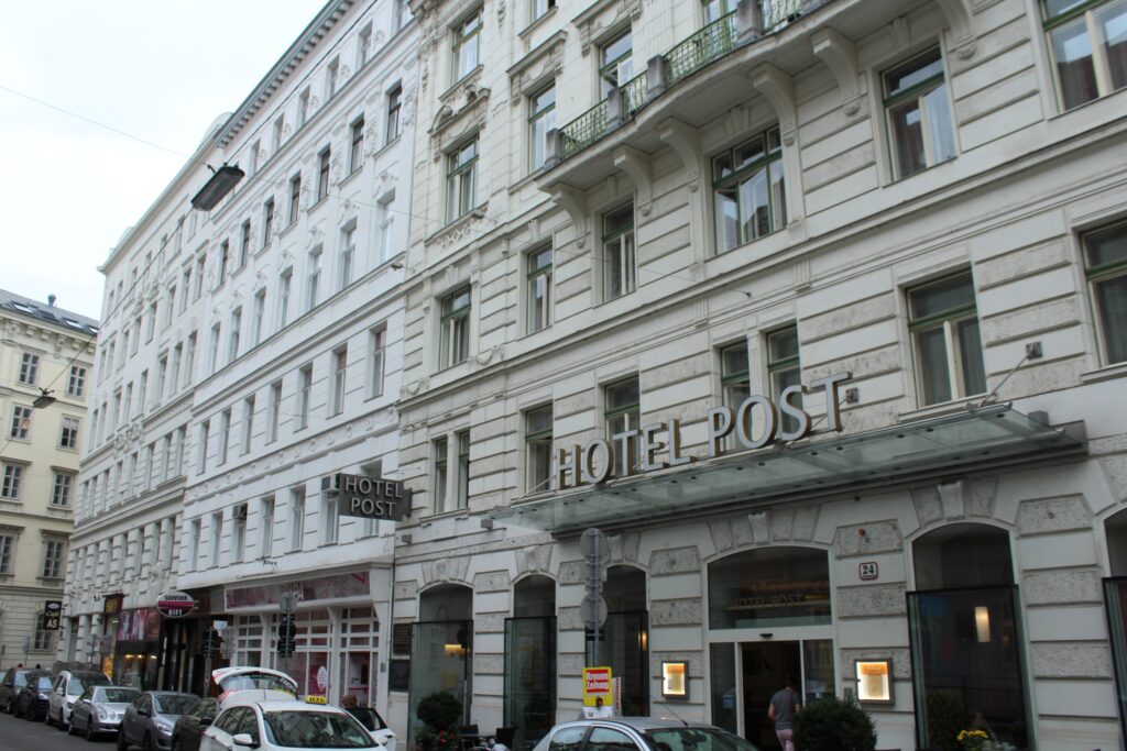 White late-historicist facade on the Fleischmarkt with iron balconies, a rusticated arched ground floor and a glass canopy with the letters HOTEL POST above number 24