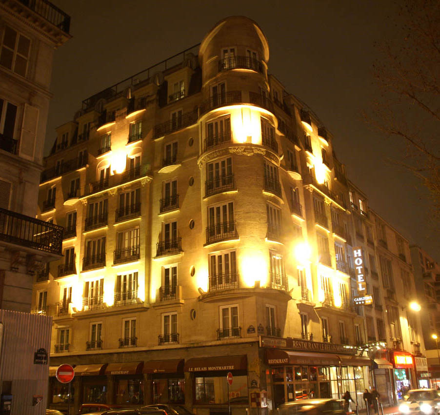 Night view of the Hôtel Rochechouart building in Paris: an eight-storey corner block in pale stone with a rounded corner rising to a small dome, iron balconies and restaurant awnings at street level