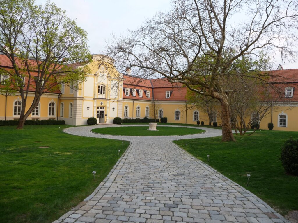Yellow Baroque manor house at Belá, Slovakia, with a white central bay under a scrolled gable, red tiled roofs with dormers and a cobbled drive round a lawn with a stone well