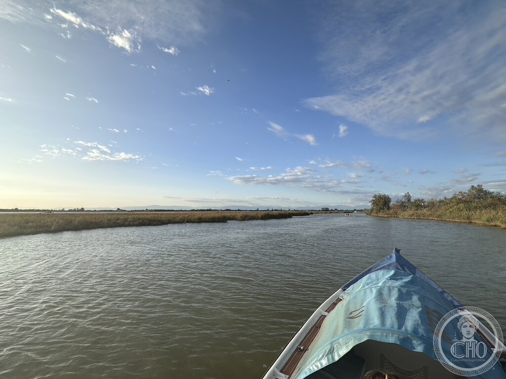 A boat navigating a narrow channel through reed beds in the Venetian Lagoon, under a wide sky