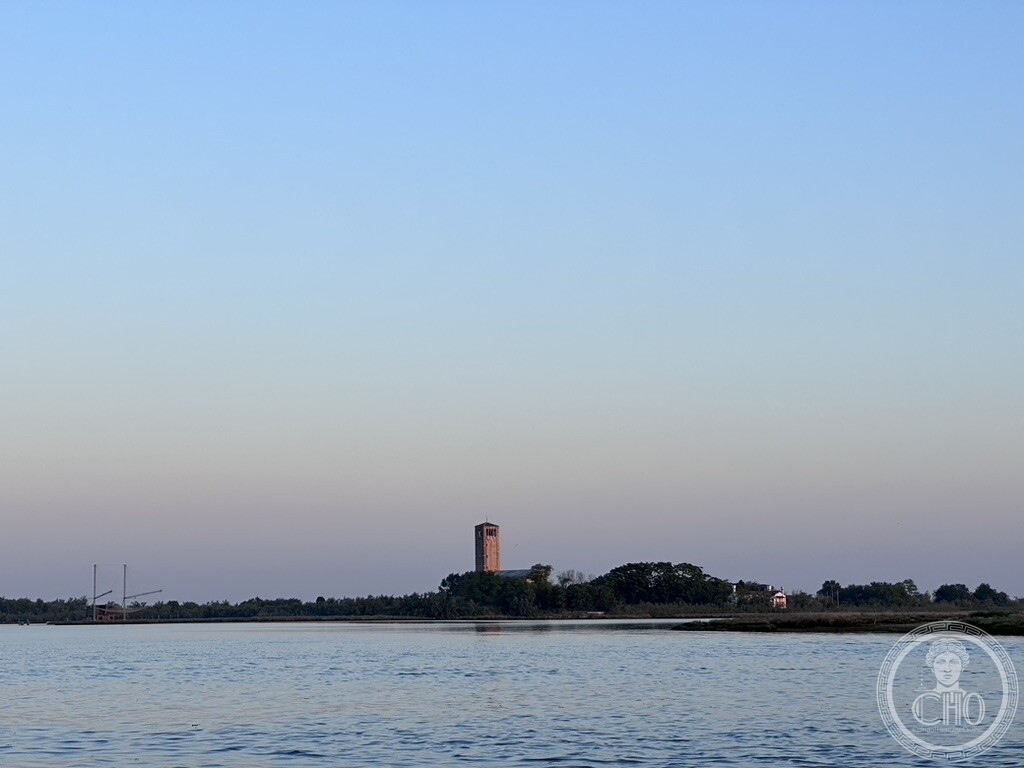The bell tower of Torcello's cathedral seen across open water at dusk, from a boat in the Venetian Lagoon