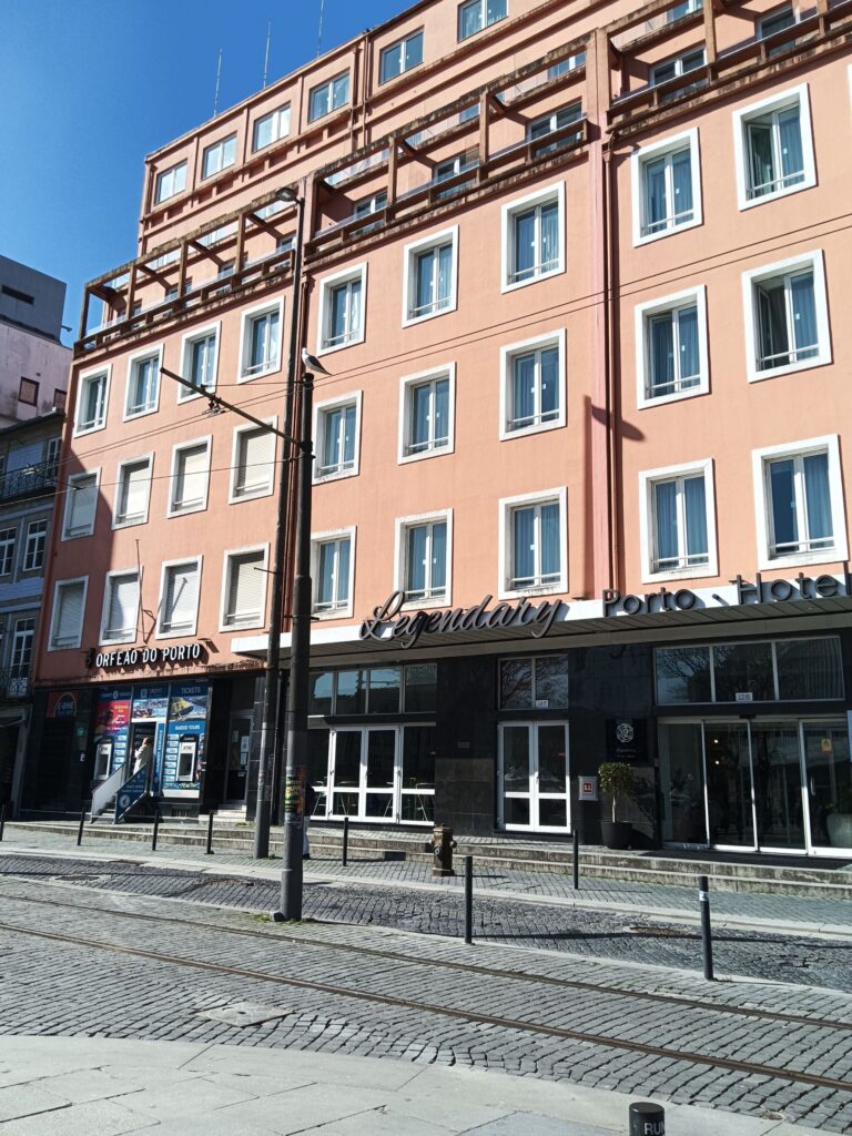 The long rendered front of the Legendary Porto Hotel on Praça da Batalha, with rows of white-framed square windows, a band of balconies with projecting brackets near the top, and a dark stone ground floor with the hotel's lettering and the sign of the Orfeão do Porto