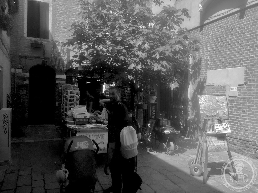 Black and white photograph of the secluded courtyard outside Libreria Acqua Alta in Venice, featuring a leafy fig tree, book barrows, print displays, and the Attenti al Gatto sign