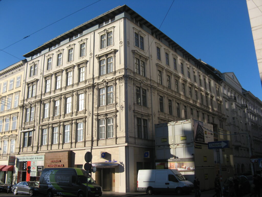 Corner house at Kärntner Straße 44 and Elisabethstraße 1 in Vienna: four storeys of pale render with paired windows under carved pediments and medallions, a projecting corner bay, modern shopfronts and the hotel entrance at street level