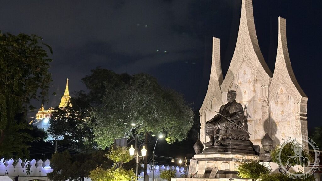 The seated bronze monument to King Rama III at night, backed by a white screen with three flame-shaped finials, with the lit golden stupa of Wat Saket beyond the trees
