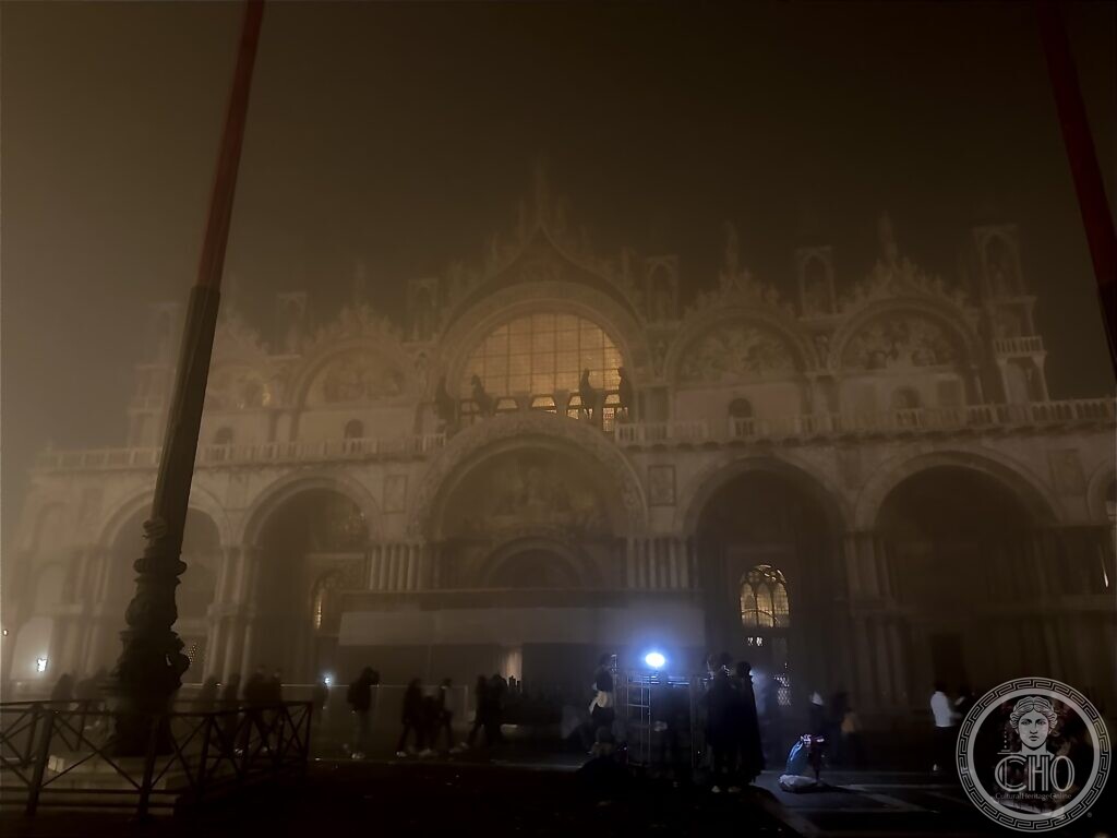 St Mark's Basilica in thick fog at night, its front glowing faintly behind a flagpole, people crossing the square