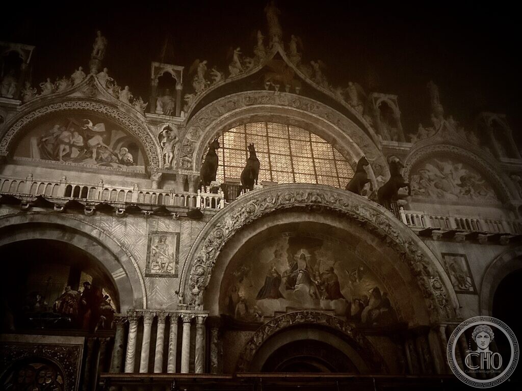 The upper front of St Mark's Basilica at night, the four bronze horses on the terrace between the arches and the mosaics of the lunettes below