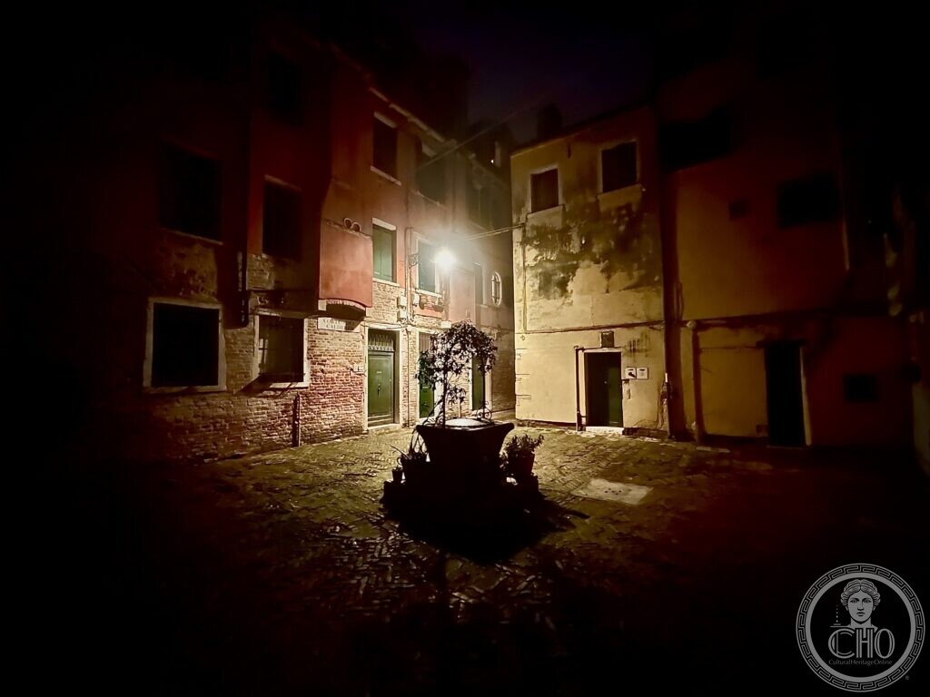 Corte del Calderer at night, the well-head with its potted tree lit by a lamp, plastered house fronts behind