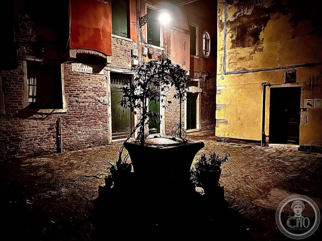 Corte del Calderer in Venice at night, a well-head topped with a potted tree at the centre, brick and plastered house fronts behind and the street sign Corte del Calderer on the corner