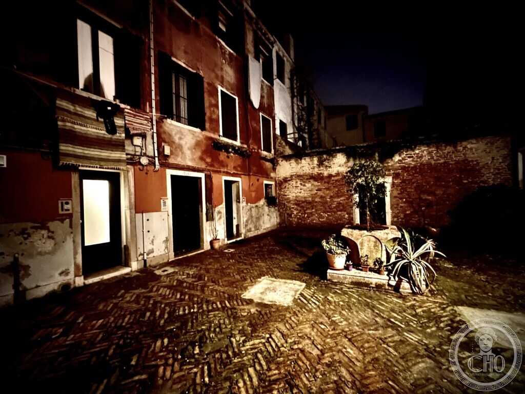 The herringbone brick paving of Corte del Calderer at night, the well-head with plants and a red house front behind