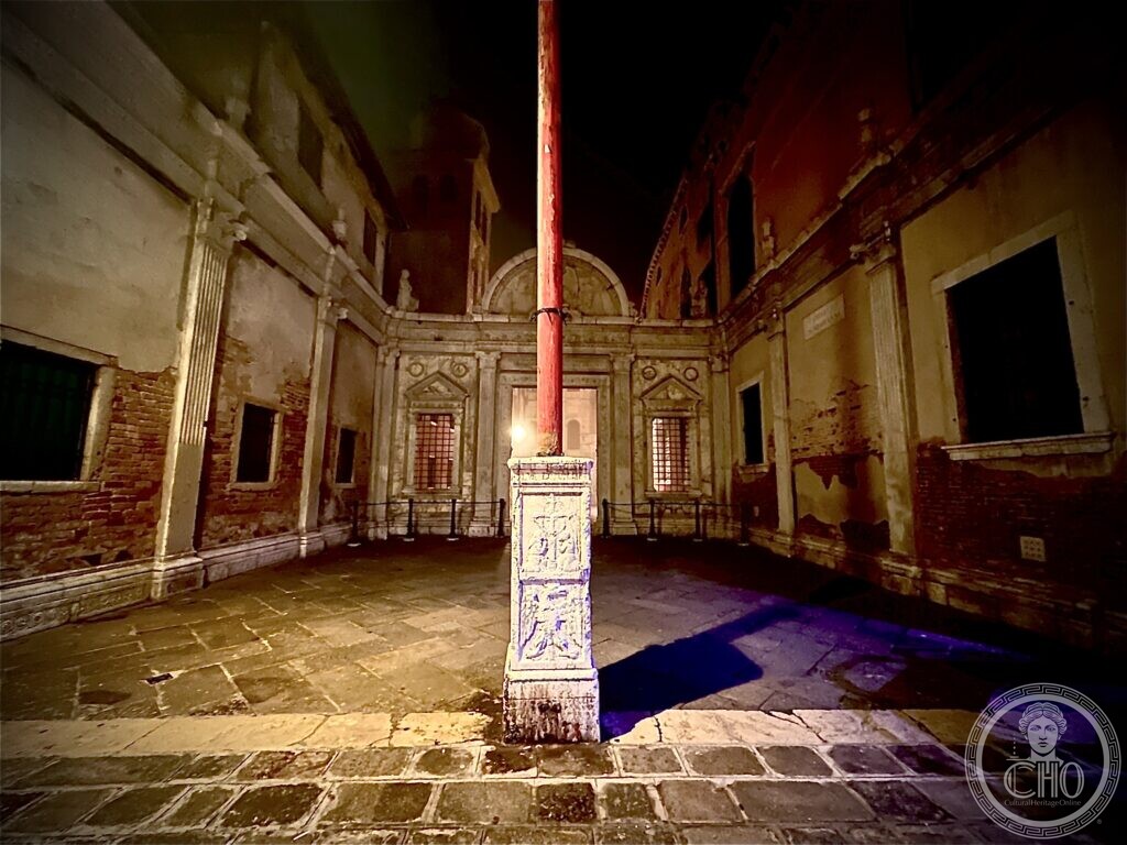 The courtyard of San Giovanni Evangelista at night, a red flagpole on a carved stone base in the foreground and Renaissance windows on the far wall
