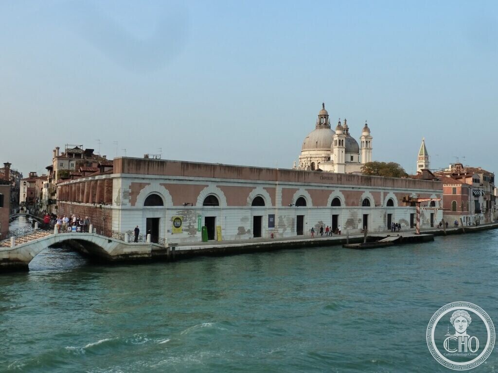 A long low warehouse front with a row of white arched openings on the Giudecca Canal, the dome of the Salute behind it and a small stone bridge on the left